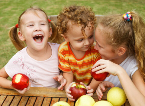 three happy children sitting at the table and eat apples
